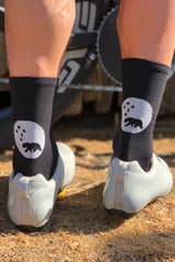 Back view Flagship Socks showing white bear logo. Model wearing black socks in white cycling shoes.